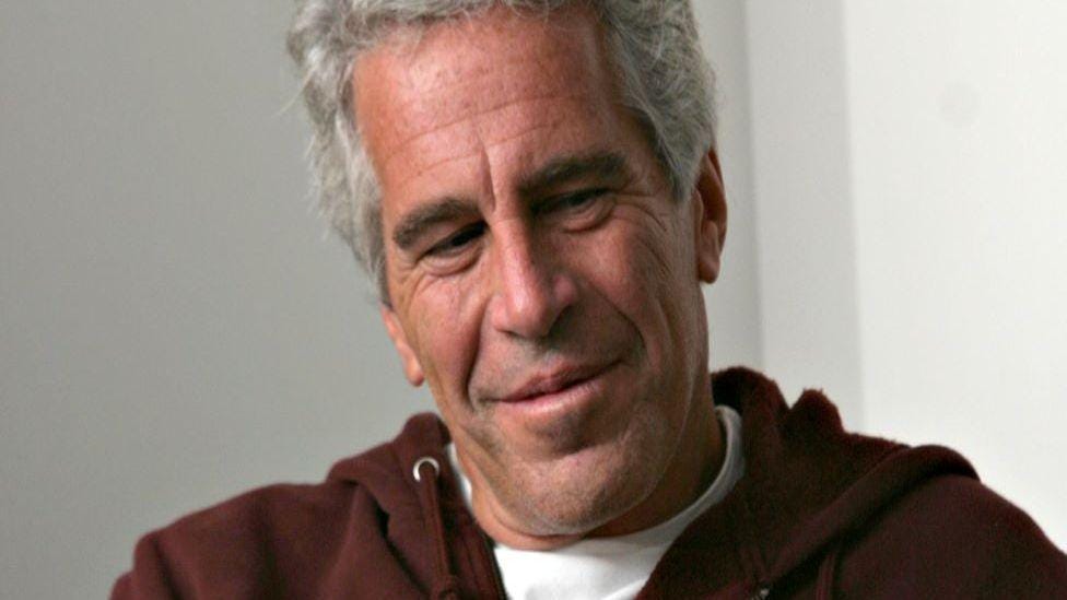 Epstein is wearing a brown hooded top with a white T shirt underneath, while his eyes are directed off to the right and there is a smile on his face.