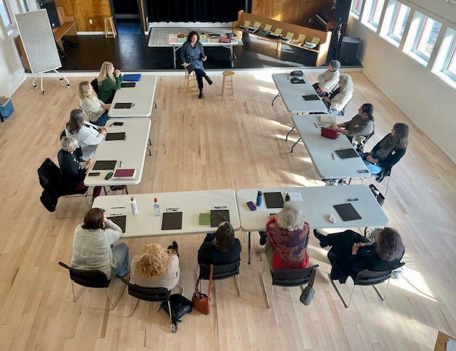 An overhead shot of a dozen people at tables in a U shape around an instructor. A row ot typewriters sits in the background