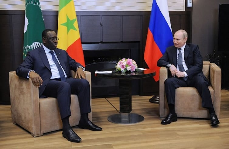Two men talk while sitting on couches in front of the flags of Russia and Senegal. Two men talk while sitting on couches in front of the flags of Russia and Senegal.