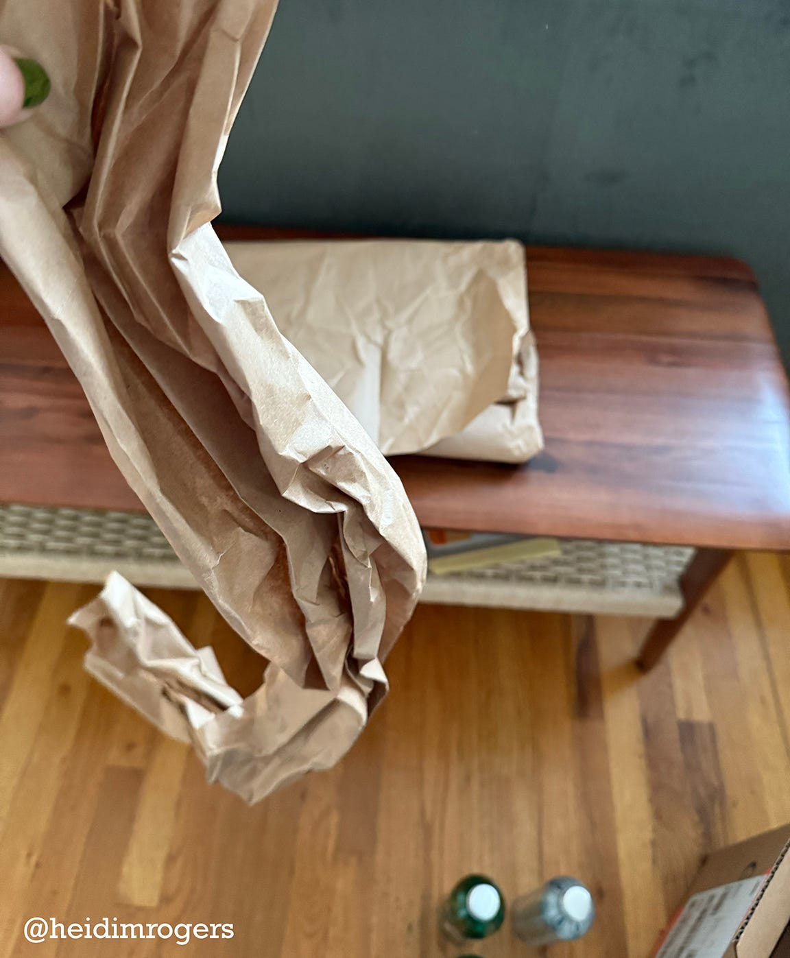 A crinkled piece of brown paper is held up in the foreground, while a folded stack of brown paper rests on top of a wooden bench in the background. In the lower right corner are two bottles of a soap and an open cardboard box.
