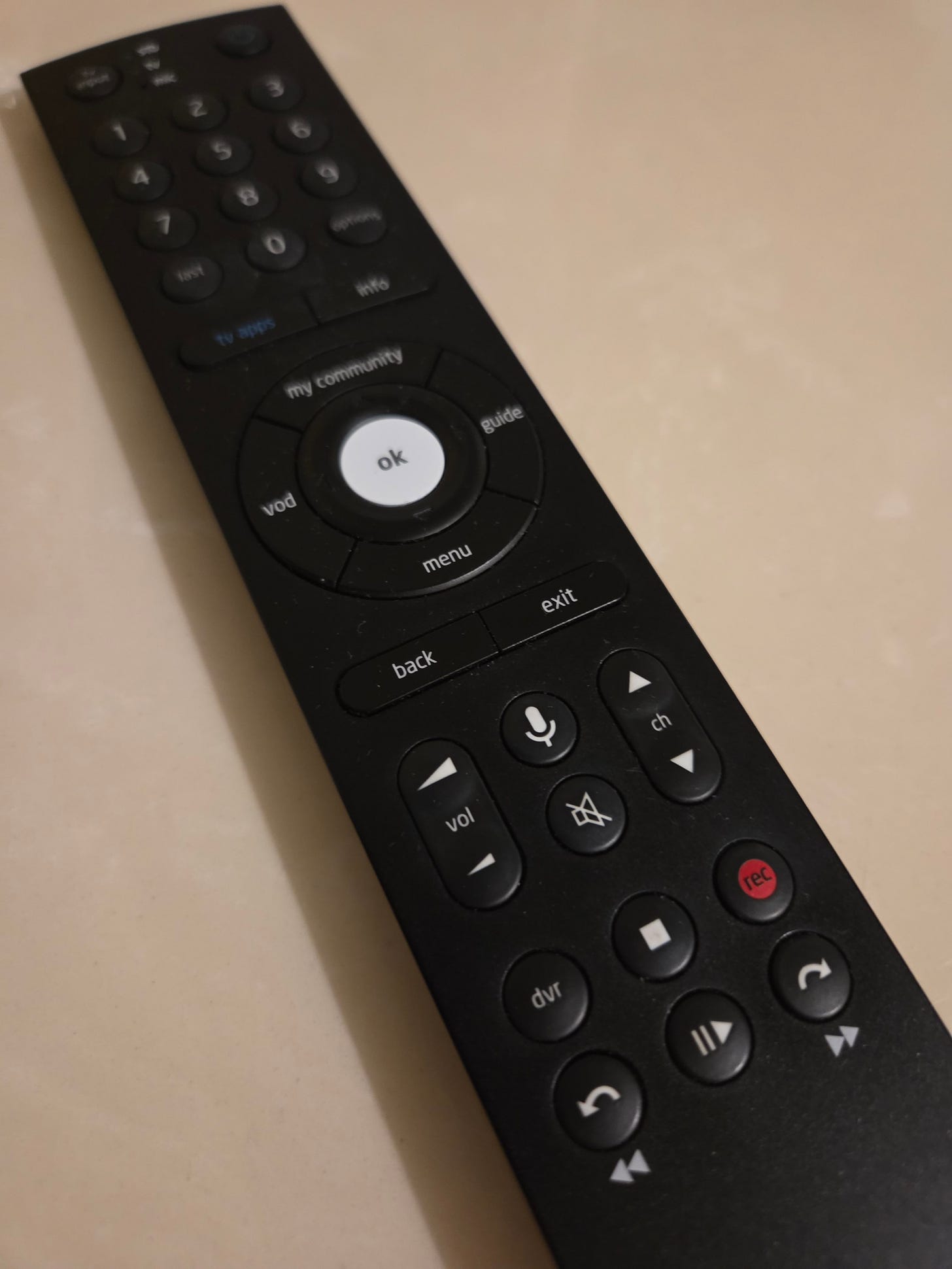 A black, plastic television remote control that is long and narrow. It has many buttons on it, including numbers, power, channel up and down, volume, and the typical controls for a DVR or VCR such as play, stop, rewind, fast forward, pause, and record.