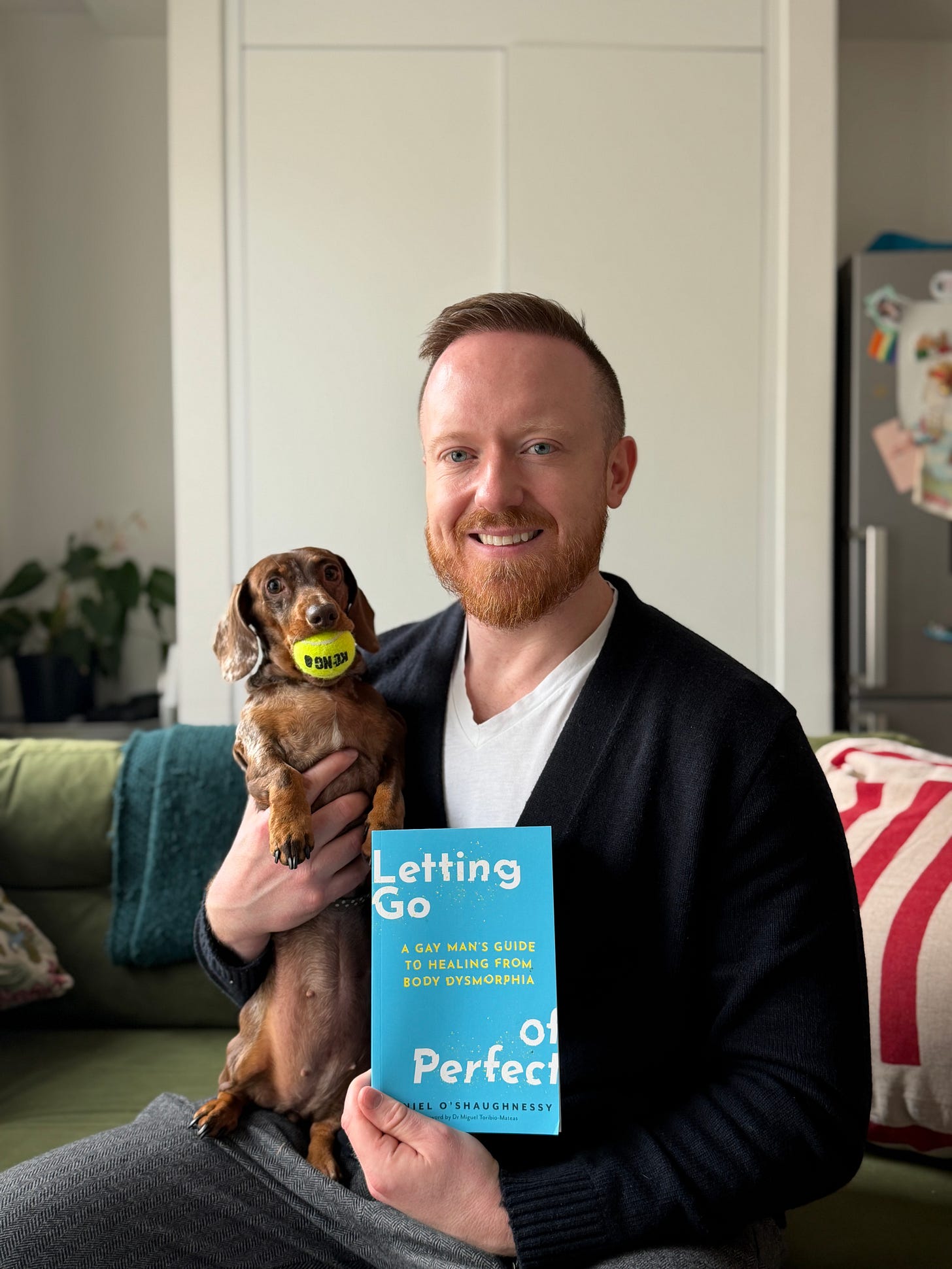 A person sitting on a sofa holding a copy of Letting Go of Perfect: A Gay Man’s Guide to Healing from Body Dysmorphia while a small brown dachshund rests on their lap.