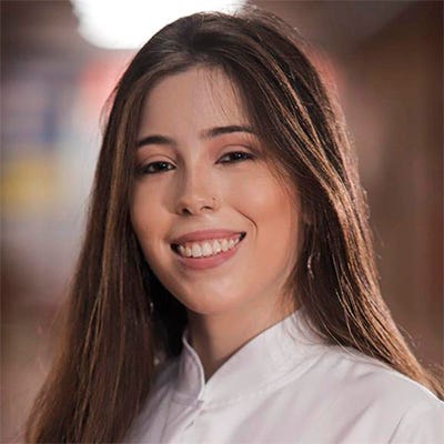 Fotografia de uma mulher jovem em um ambiente interno, sorrindo para a câmera. Ela tem cabelos longos e castanhos e usa uma roupa profissional branca. No bolso da roupa há uma identificação com o nome “Dra. Anna Luísa A. Pimenta” e a indicação “Cirurgiã-dentista”. Ao fundo, há um corredor ou ambiente institucional desfocado. Fotografia de uma mulher jovem em um ambiente interno, sorrindo para a câmera. Ela tem cabelos longos e castanhos e usa uma roupa profissional branca. No bolso da roupa há uma identificação com o nome “Dra. Anna Luísa A. Pimenta” e a indicação “Cirurgiã-dentista”. Ao fundo, há um corredor ou ambiente institucional desfocado.
