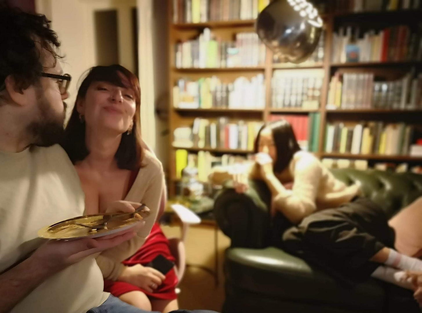 Interno di una casa piena di libri. Elena, una donna sui 40 anni, sorridente e piena di sfida, guarda in camera. Intorno a lei, persone che mangiano e si rilassano. Interno di una casa piena di libri. Elena, una donna sui 40 anni, sorridente e piena di sfida, guarda in camera. Intorno a lei, persone che mangiano e si rilassano.