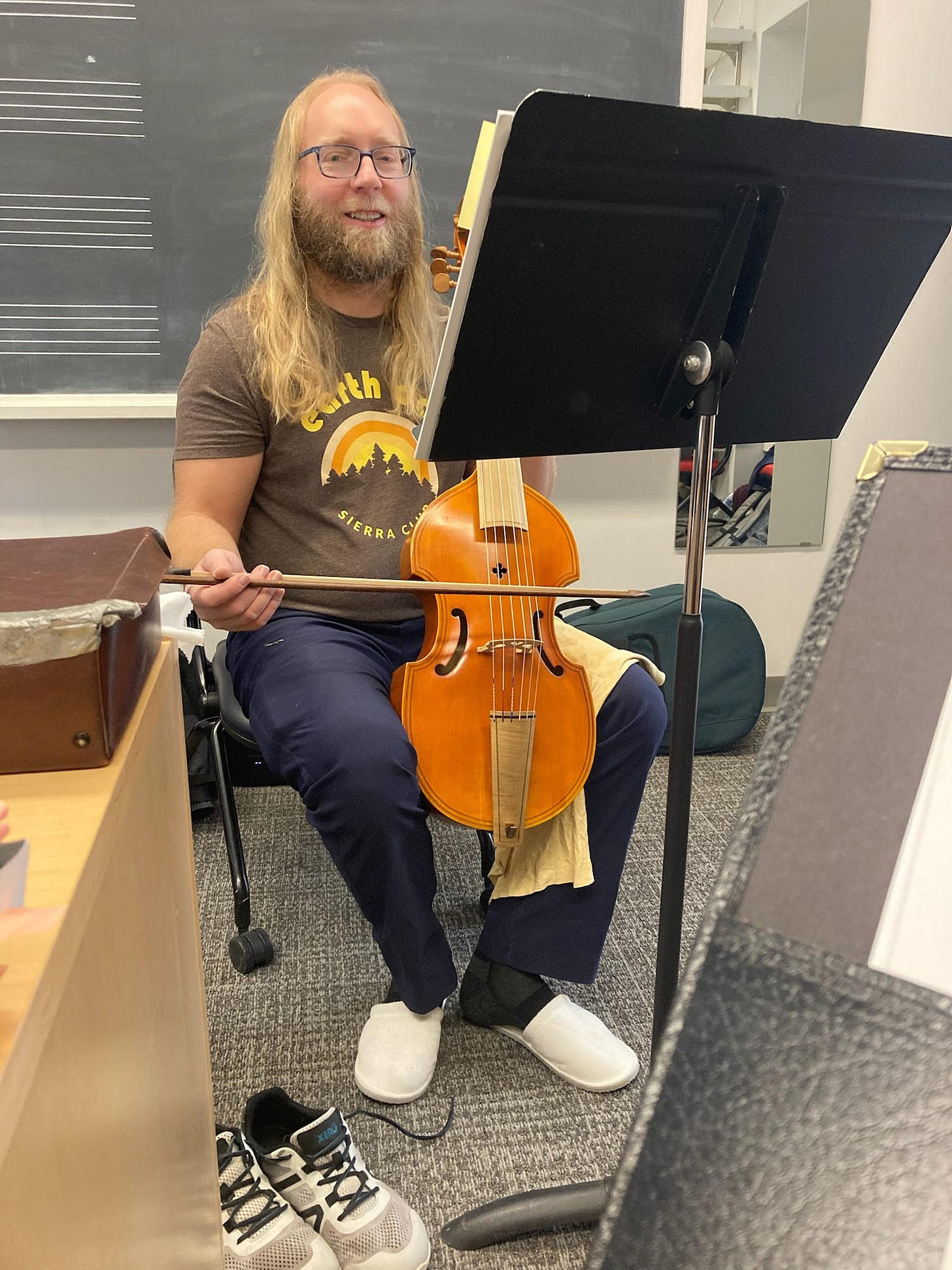 Author with tenor viol held between kness, holding bow ready to play. Smiling blond man with beard and long hair, and glasses.
