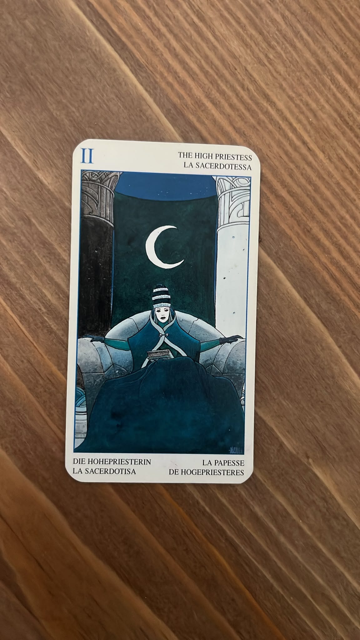 The High Priestess tarot card lies on a wooden table, showing a seated woman in dark blue beneath a crescent moon between black and white pillars. The High Priestess tarot card lies on a wooden table, showing a seated woman in dark blue beneath a crescent moon between black and white pillars.