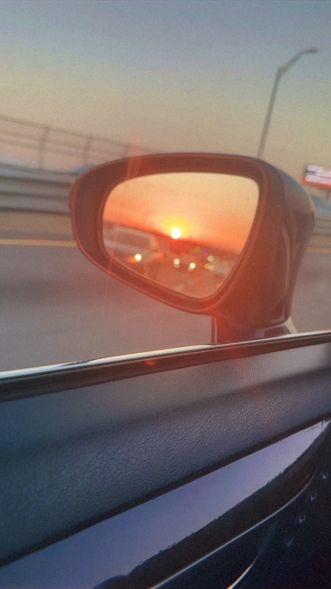 Image of a rearview mirror that shows the reflection of the sun and a peach gradient sky with cars in the forefront.