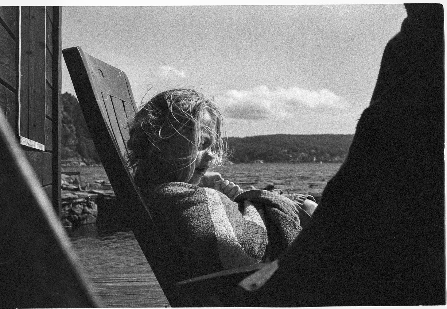 One child reclining on a chair by an ocean bay explains something to another child with serious mannerisms. Both children wear a towel. One child reclining on a chair by an ocean bay explains something to another child with serious mannerisms. Both children wear a towel.