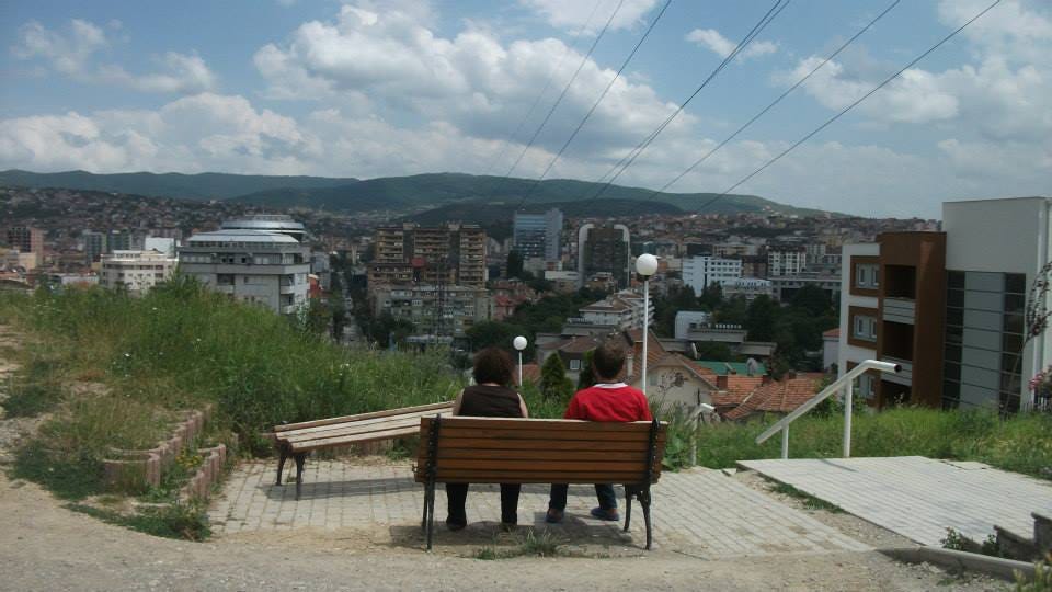 A woman and younger man sit on a wooden bench with their backs to the camera overlooking Pristina A woman and younger man sit on a wooden bench with their backs to the camera overlooking Pristina