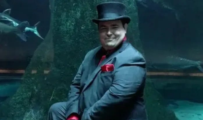 A photograph of a man in a bowler hat and suit standing in front of a shark tank.