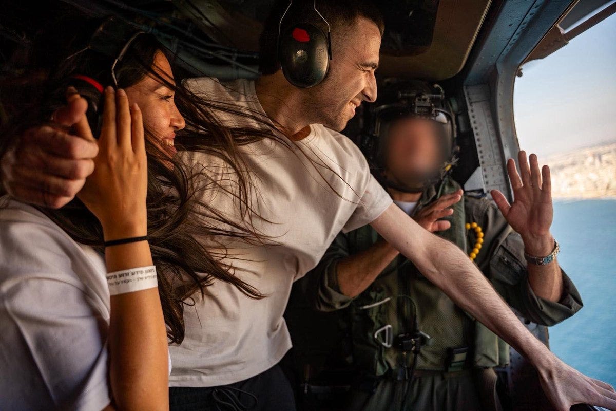 Inside a helicopter cockpit with blue sea visible through open doors, a woman with long dark hair wearing a white top and wristband hugs a man in a white t-shirt from behind while he smiles and raises his hand in a waving gesture, another figure in military uniform with blurred face extends a hand in greeting nearby.