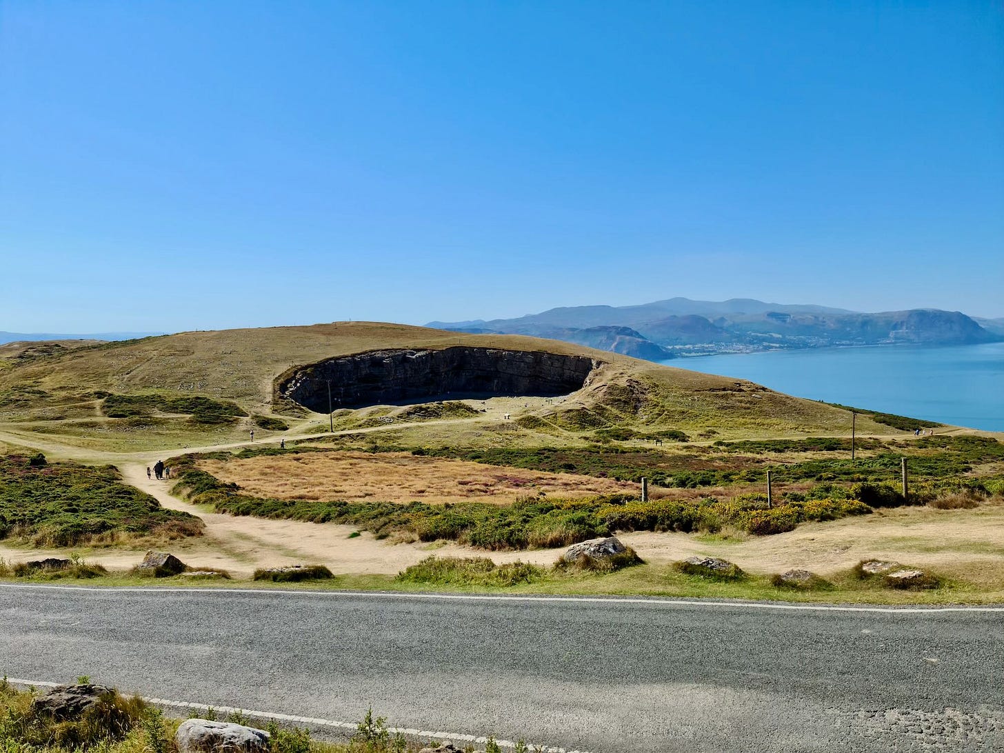 A view of Llandudno’s Great Orme: a grass-covered mountain with a distant quarry, a bay and the sea below, and a road visible from the tram. The sky is bright blue. 