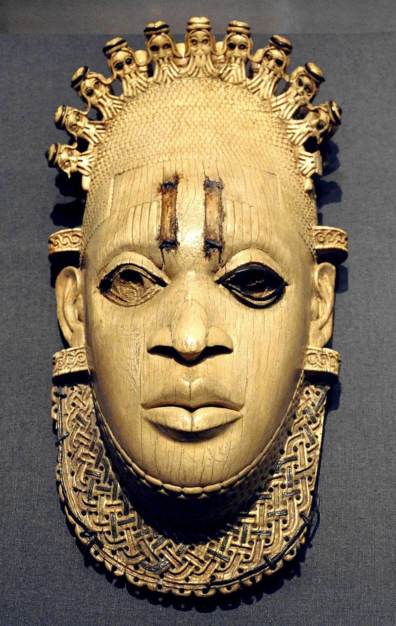 A carved African ivory mask on a dark grey background. The mask has intricate carved details around its edges depicting small figures linking arms across the crown of its head and an interwoven textural piece around the base of it jawline.