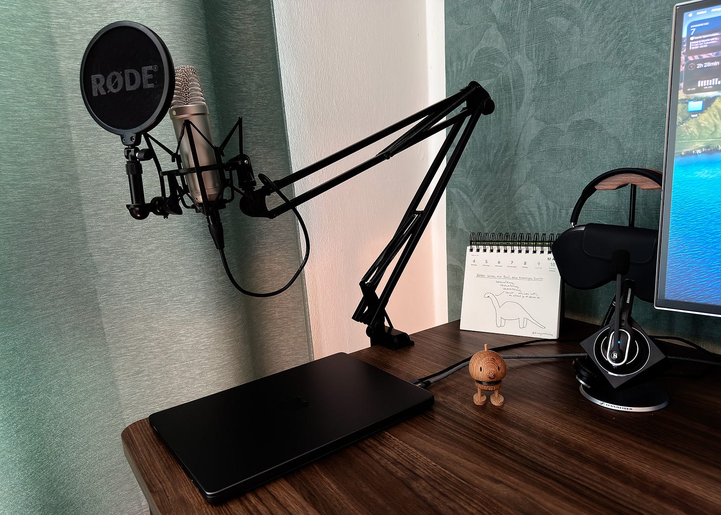 Ein Heimstudio-Schreibtisch aus dunklem Holz mit Audio-Equipment. Im Vordergrund ein Rode NT1-Kondensatormikrofon auf einem schwarzen Gelenkarm-Schwenkarm, mit Spinne und rundem Popschutz mit Rode-Logo. Auf dem Tisch liegt ein schwarzes Macbook Pro. Im Hintergrund steht ein aufgeschlagener Spiralblock-Kalender mit einer handgezeichneten Dinosaurier-Skizze und handschriftlichen Notizen. Rechts daneben ein Sennheiser-Kopfhörer auf einem Holz-Ständer sowie ein Monitor, auf dem ein Bildschirm mit einer Landschaftsansicht zu erkennen ist. Zwischen Notizbuch und Kopfhörer steht eine kleine hölzerne Hoptimist-Figur. Der Hintergrund zeigt eine grüne Tapete mit Blättermuster.