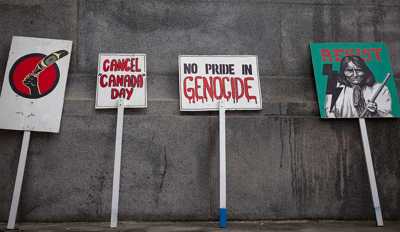 Signs reading “Cancel ‘Canada’ Day,” “No Pride in Genocide,” and “Resist,” propped against a wall. 