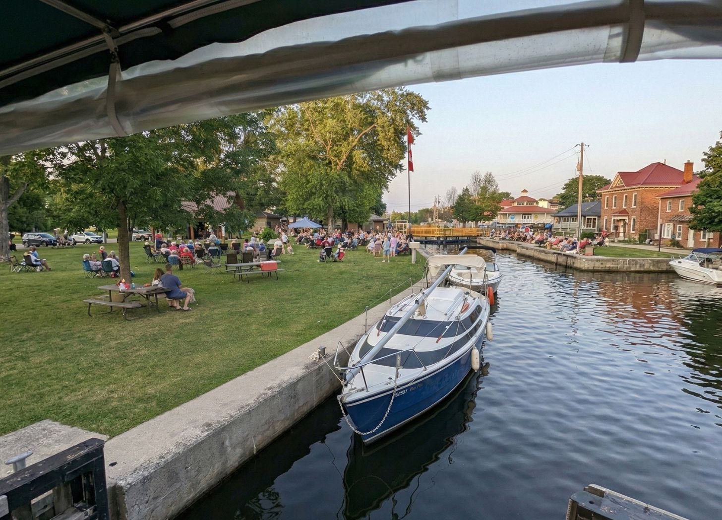 Boats passing through the lock in Bobcaygeon, Ontario, with people gathered along the canal and seated on the grassy park nearby. Boats passing through the lock in Bobcaygeon, Ontario, with people gathered along the canal and seated on the grassy park nearby.