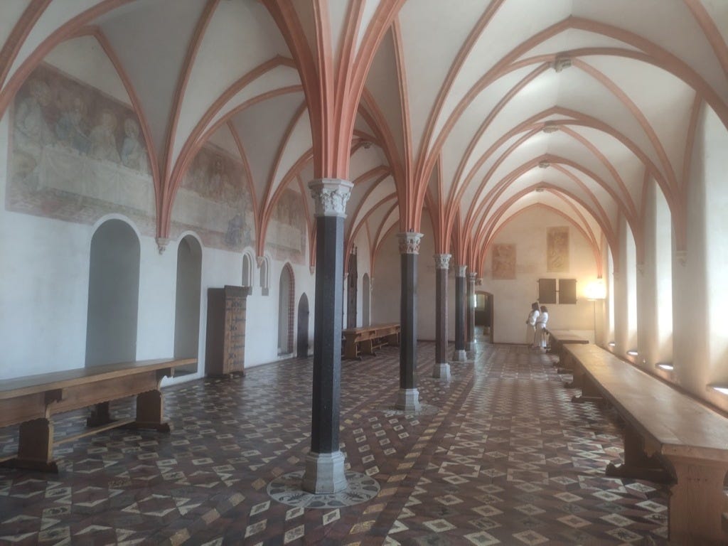 A long room, with white walls and painted frescoes above head height. Tall black pillars hold the arches that spread across the ceiling.