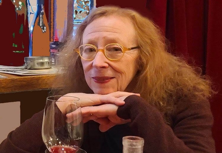 a photo of Alice Feiring smiling and posing with her hands below her chin with a glass of wine in front of her, red velvet drapes behind