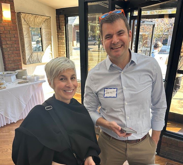 A gallery of four images. The upper left-hand image is of a white woman with short blonde hair and a black sweater shawl standing next to a tall white man with brown hair and wearing a light blue button-down, with orange sunglasses perched on his head. They are standing in a well-lit lobby in front of a wall of windows and a table with a white tablecloth. The upper right picture is of a very diverse group of people sitting at tables with white tablecloths in a well-lit lobby. There are drinks, flowers, and napkins on the tables. A Black woman stands at the front of the room holding a microphone. The lower left-hand picture shows a white man with mid-tone brown hair and a navy blue sweater with blue plaid shirt underneath in the foreground. He is seated at a table with a white tablecloth with flowers, plates of food, and drinks on it. He is surrounded by a diverse group of people all attentively facing the front of the room to the left. The bottom right-hand image is a zoomed-out image of another table with white tablecloth, food, and drinks in the same well-lit lobby with a wall of glass windows. A diverse group of people sits at the table and is looking away from the front of the room, presumably at someone at another table.