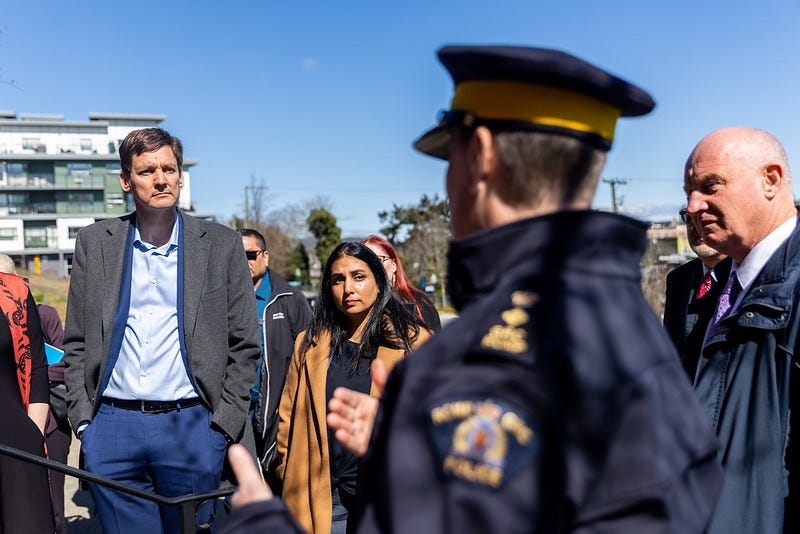 premier david eby stands with his hands in his pockets next to attorney general niki sharma, who also has her hands in her pockets. to the right is solicitor general and public safety minister mike farnworth. all are looking with serious expressions at an RCMP officer who is blurry in the foreground