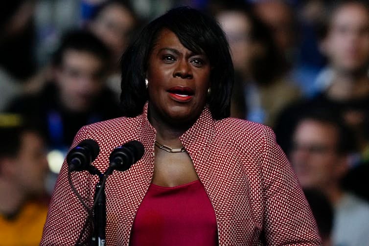Woman in red shirt and red and white patterned jacket speaks in front of microphone to an audience Woman in red shirt and red and white patterned jacket speaks in front of microphone to an audience