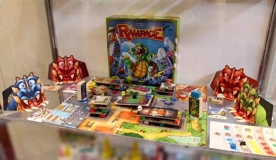 The board game Rampage set out on a table. Meeple hold up buildings in a cardboard city while wooden dinosaurs stand on the colorful board.