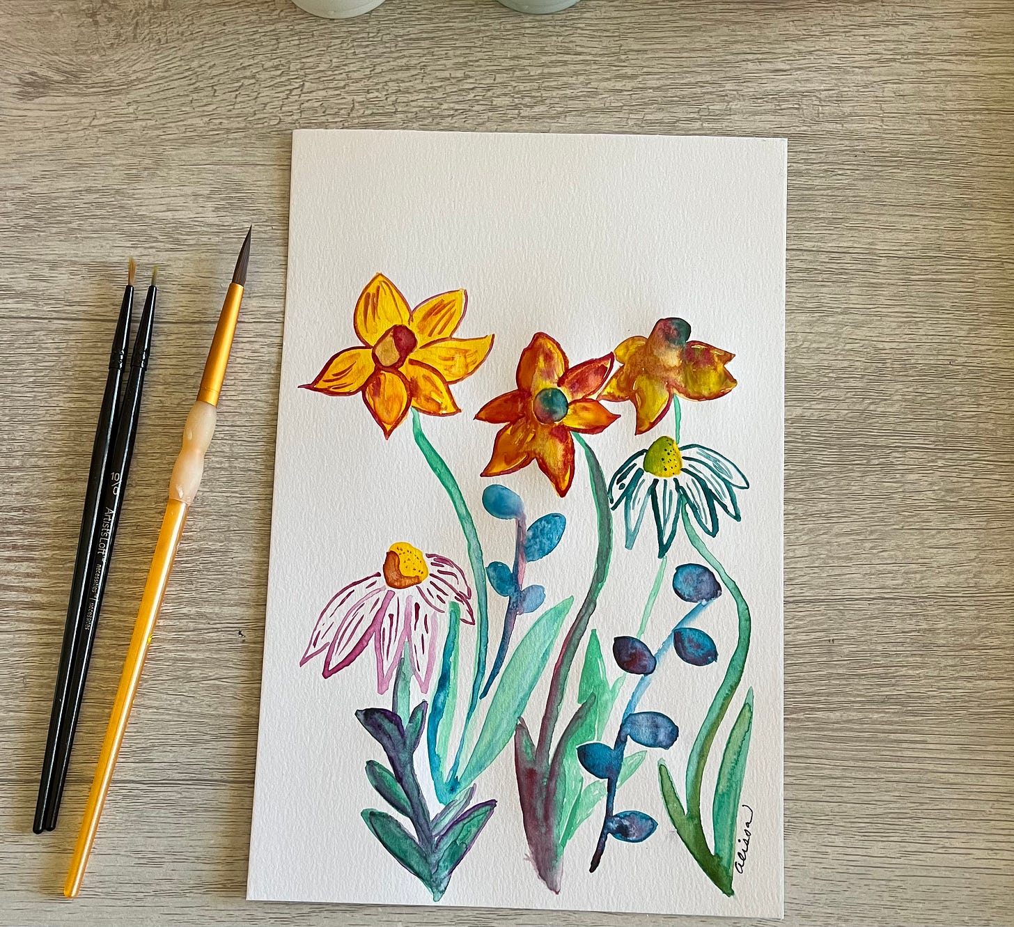 Photo of a watercolor and gauche painting of some daffodils and other flowers on a desk beside some paintbrushes. Photo of a watercolor and gauche painting of some daffodils and other flowers on a desk beside some paintbrushes.