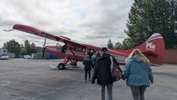 people walking toward a small red airplane on a small airstrip under a mostly cloudy sky