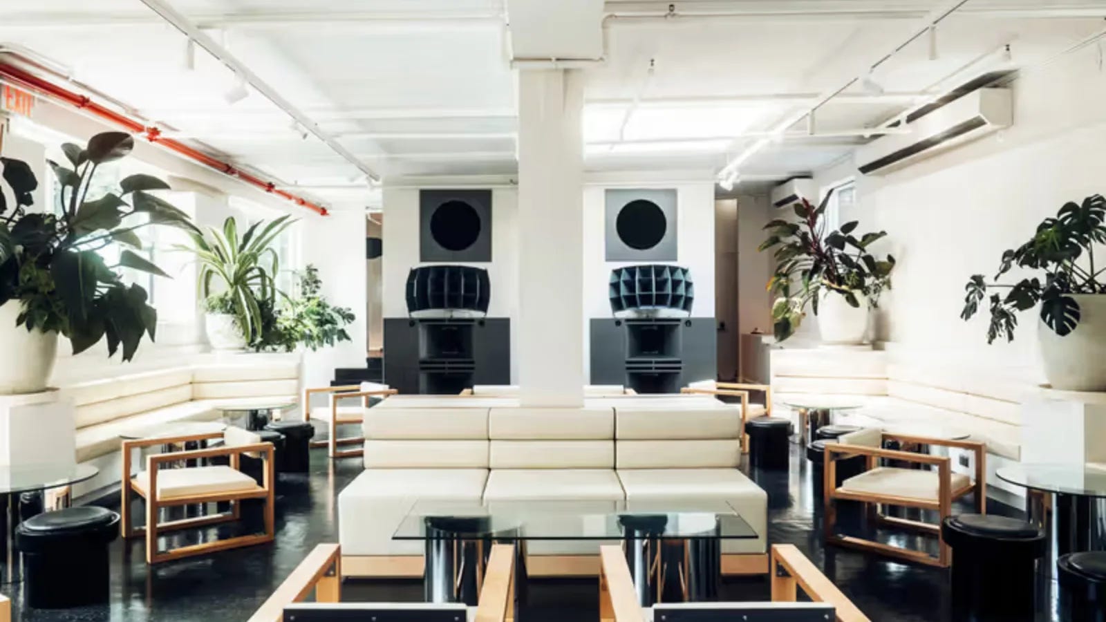 The daylit room at Public Records: cream modular seating and blond-wood chairs on a black gloss floor, big potted plants along the windows, and two black horn speakers flanking the central column