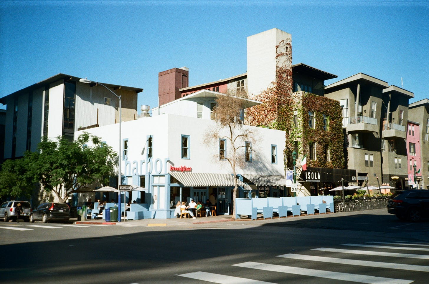 Early morning photo of Harbor Breakfast, San Diego. The entire diner can be seen with the crosswalk in the foreground. Sky blue furniture and striped parasols line the outside walls of the building.