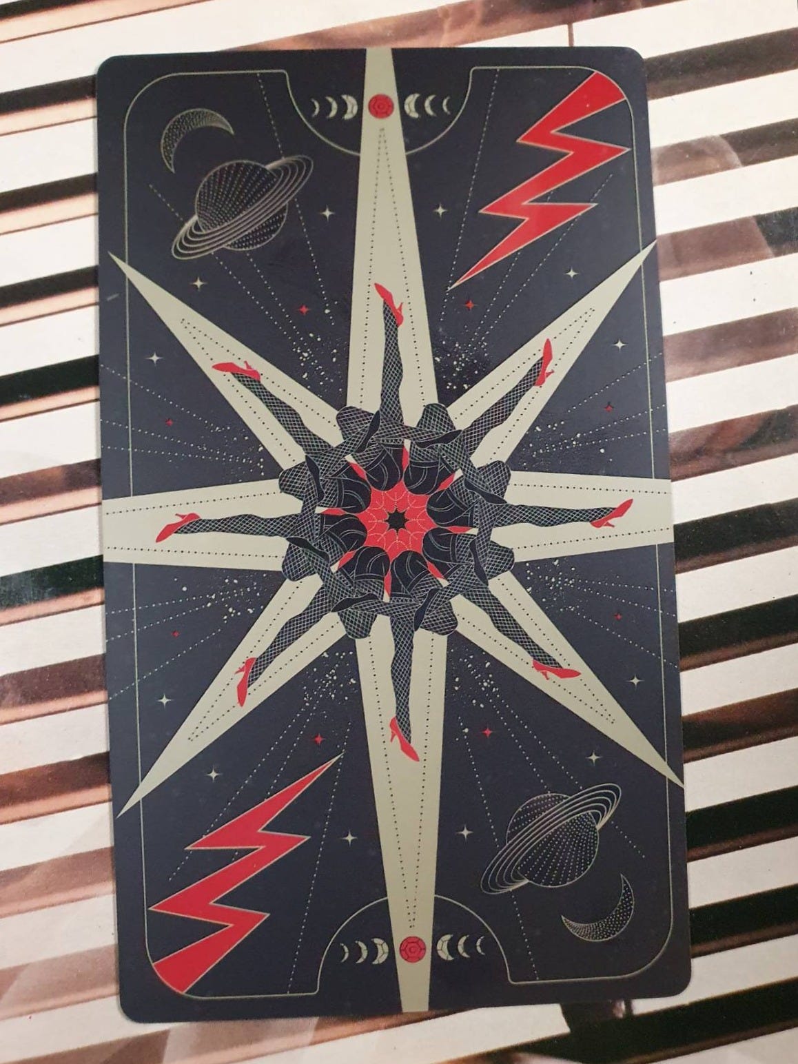 An elaborate tarot card back featuring a symmetrical circle of black-stockinged legs in red heels, surrounded by celestial symbols, crescent moons, planets and red lightning bolts. An elaborate tarot card back featuring a symmetrical circle of black-stockinged legs in red heels, surrounded by celestial symbols, crescent moons, planets and red lightning bolts.