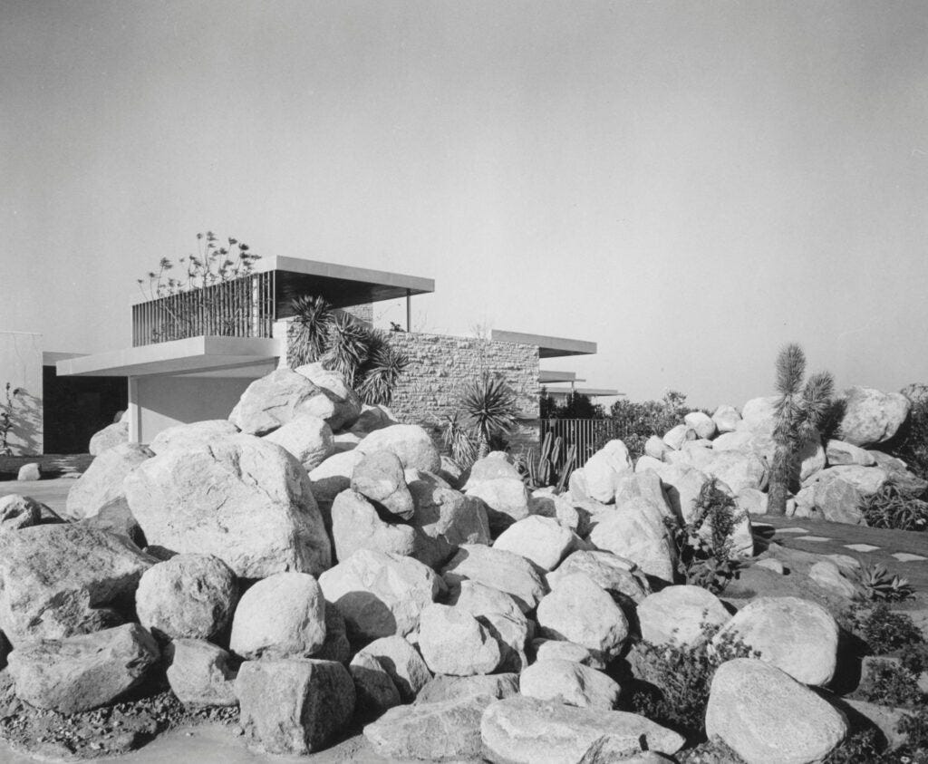 A modern architectural structure surrounded by large boulders and desert plants, captured in black and white.