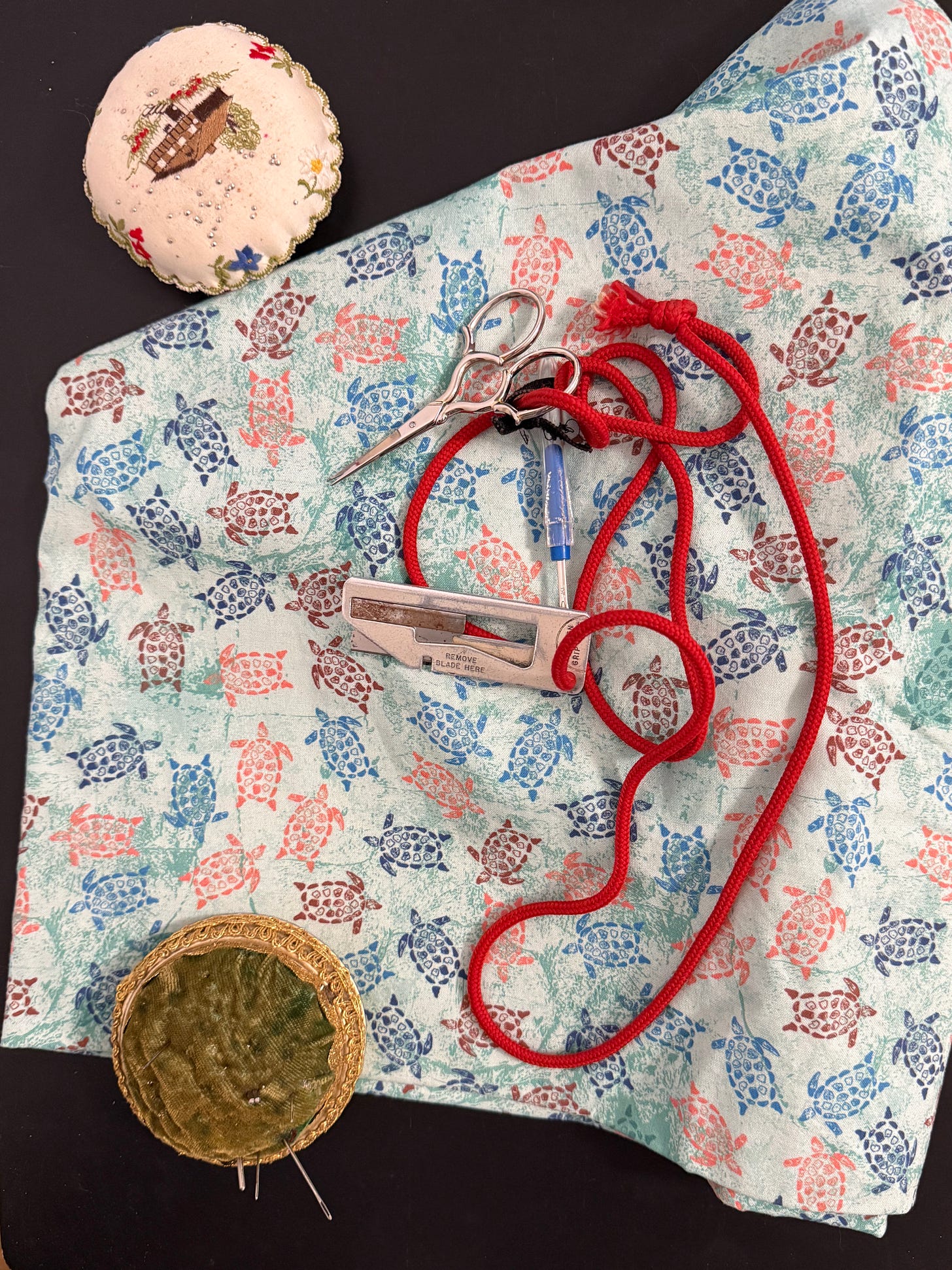Chatelaine with open razor, embroidery scissors, and seam ripper arranged on light blue sea turtle print fabric with two pin cushions. Chatelaine with open razor, embroidery scissors, and seam ripper arranged on light blue sea turtle print fabric with two pin cushions.