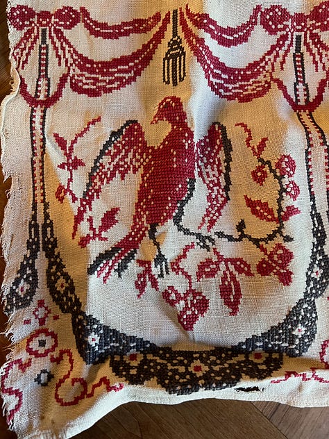Three photos of embroidery work. Left, a white linen tablecloth edged with lace and embroidered with a geometric pattern in red; in the middle a kind of cotton net sewn with decorative patterns in red, yellow, and green yarn with "1906" in the middle; on the right, a white linen table runner embroidered with a red bird and black decorative edging.