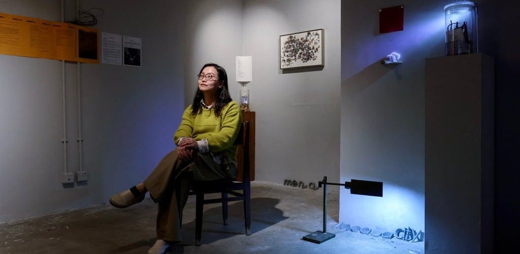 A woman sits in a chair inside a room in an art gallery with various kinds of lighting on artworks hanging on walls and resting on the floor.