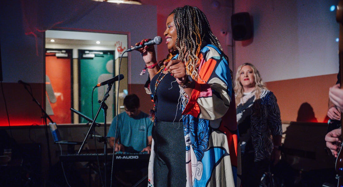 A woman - Davina Songbird - wearing a colourful cloak singing into a microphone, with a band around her and colourful lights