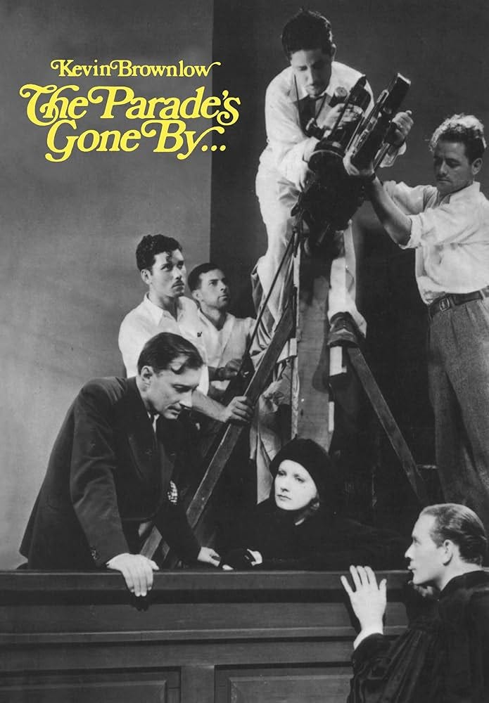 A black and white photo of a cameraman in the 1920s filming a scene with Greta Garbo. Crew surround them. In yellow on the top left corner reads Kevin Brownlow and underneath, The Parade’s Gone By. A black and white photo of a cameraman in the 1920s filming a scene with Greta Garbo. Crew surround them. In yellow on the top left corner reads Kevin Brownlow and underneath, The Parade’s Gone By.