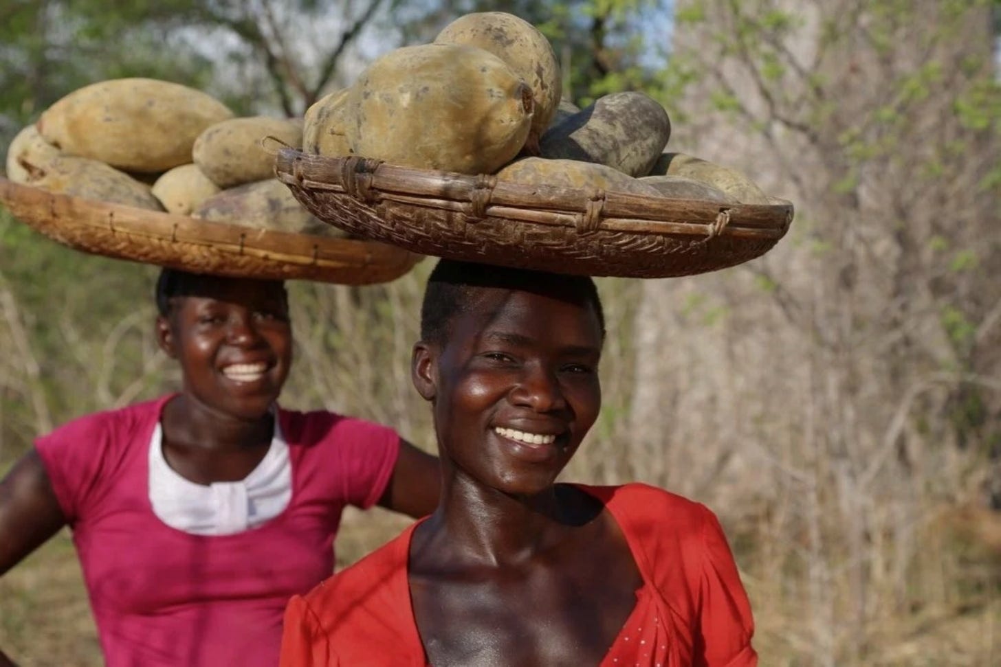 Two women in red dresses smiling and carrying baskets of shea nuts