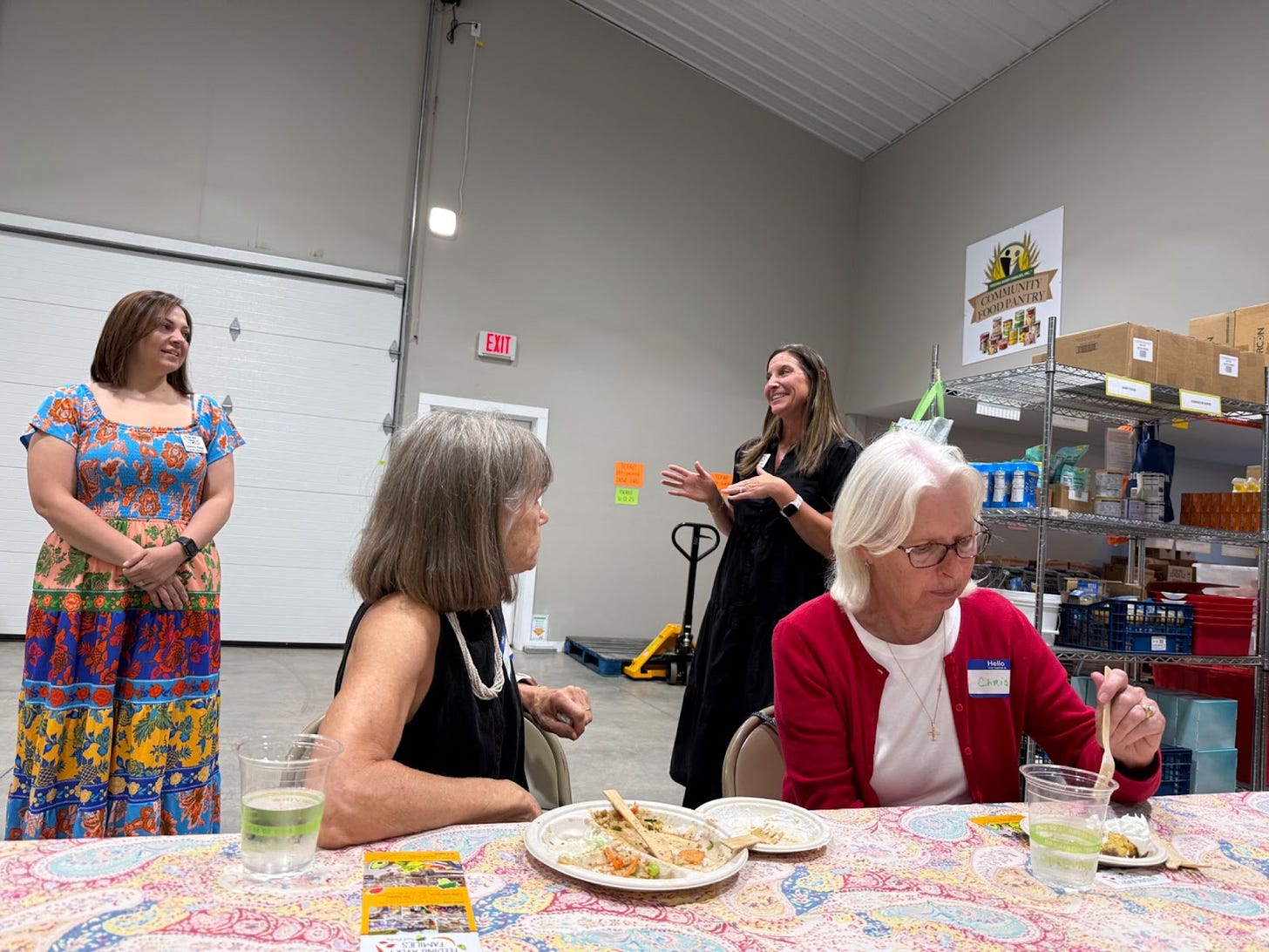 Women gathered around tables in a food pantry warehouse, sharing a potluck meal with volunteers and food distribution shelves visible in the background. Women gathered around tables in a food pantry warehouse, sharing a potluck meal with volunteers and food distribution shelves visible in the background.