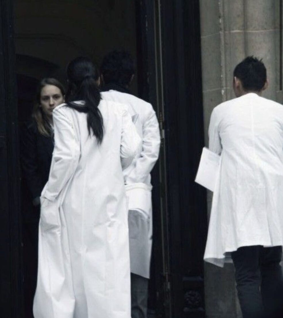 “Photographic and archival imagery of individuals wearing white lab coats as used in the design studio of Maison Martin Margiela. These garments functioned as a symbolic uniform to neutralize hierarchy, promote anonymity, and emphasize collective authorship over individual style. The white coats reflect the psychological and symbolic aspects of enclothed cognition — clothing as a trigger for behavior, identity, and group dynamics.”