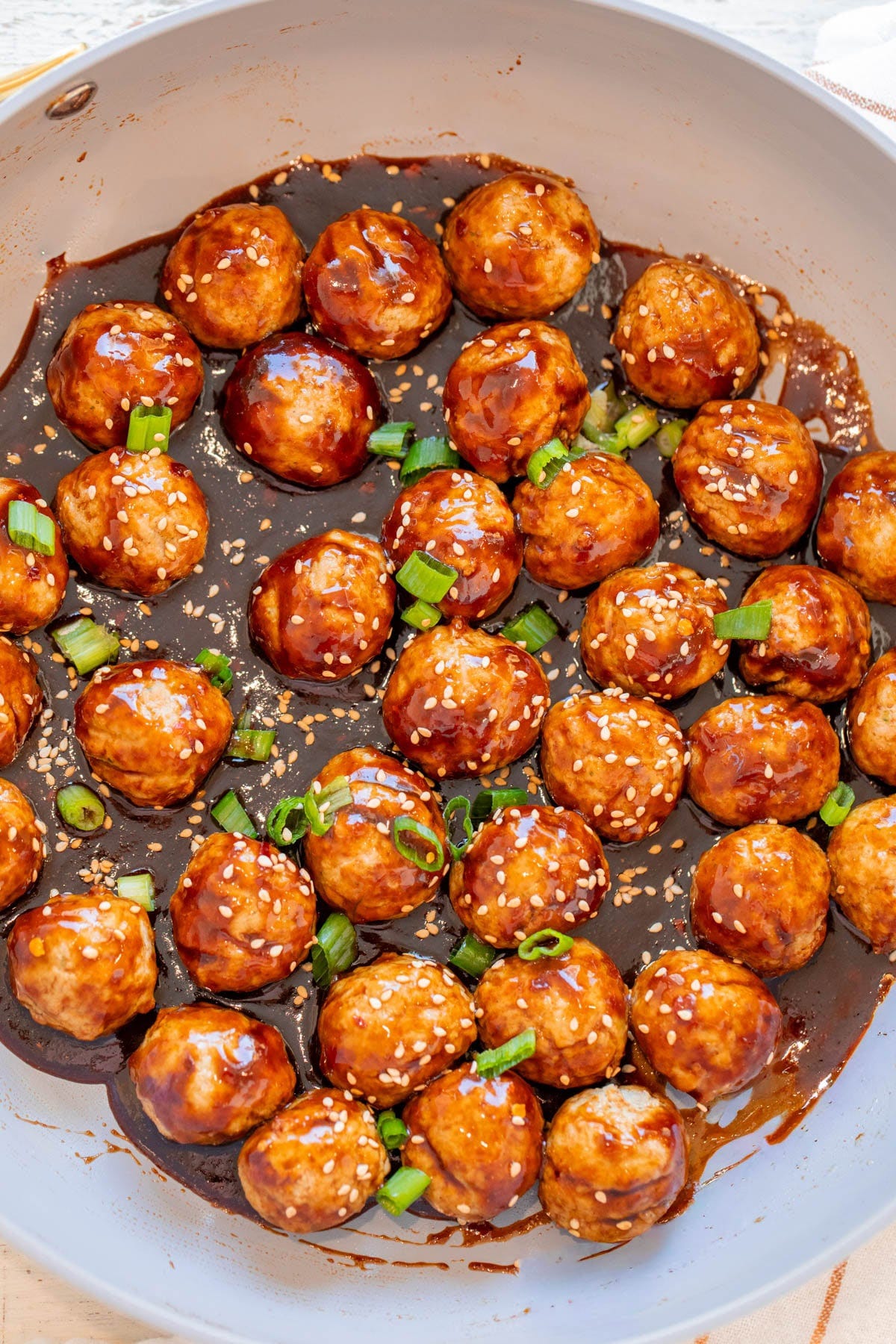 Meatballs coated in a dark glaze, garnished with sesame seeds and chopped green onions, arranged in a pan.