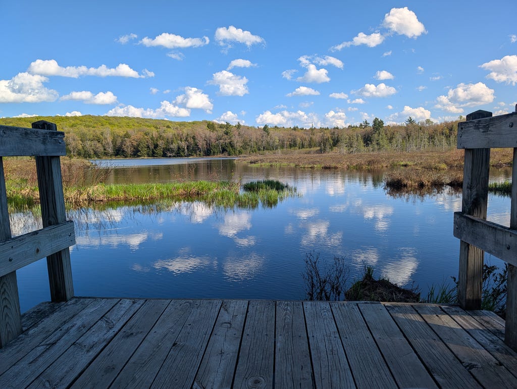 Small puffy clouds in a blue sky are reflected on the surface of a small pond, with green hills in the distance and a small dock visible in the foreground. Small puffy clouds in a blue sky are reflected on the surface of a small pond, with green hills in the distance and a small dock visible in the foreground.
