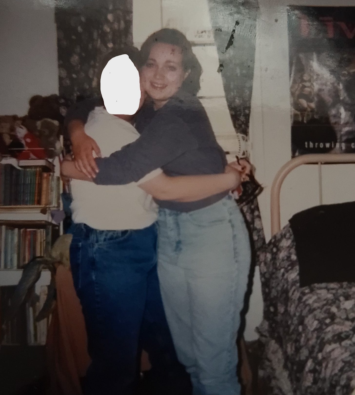 Two caucasian teen girls embrace in a 1990's bedroom, complete with a poster from the band LIVE and a flowered bedspread on the bed.
