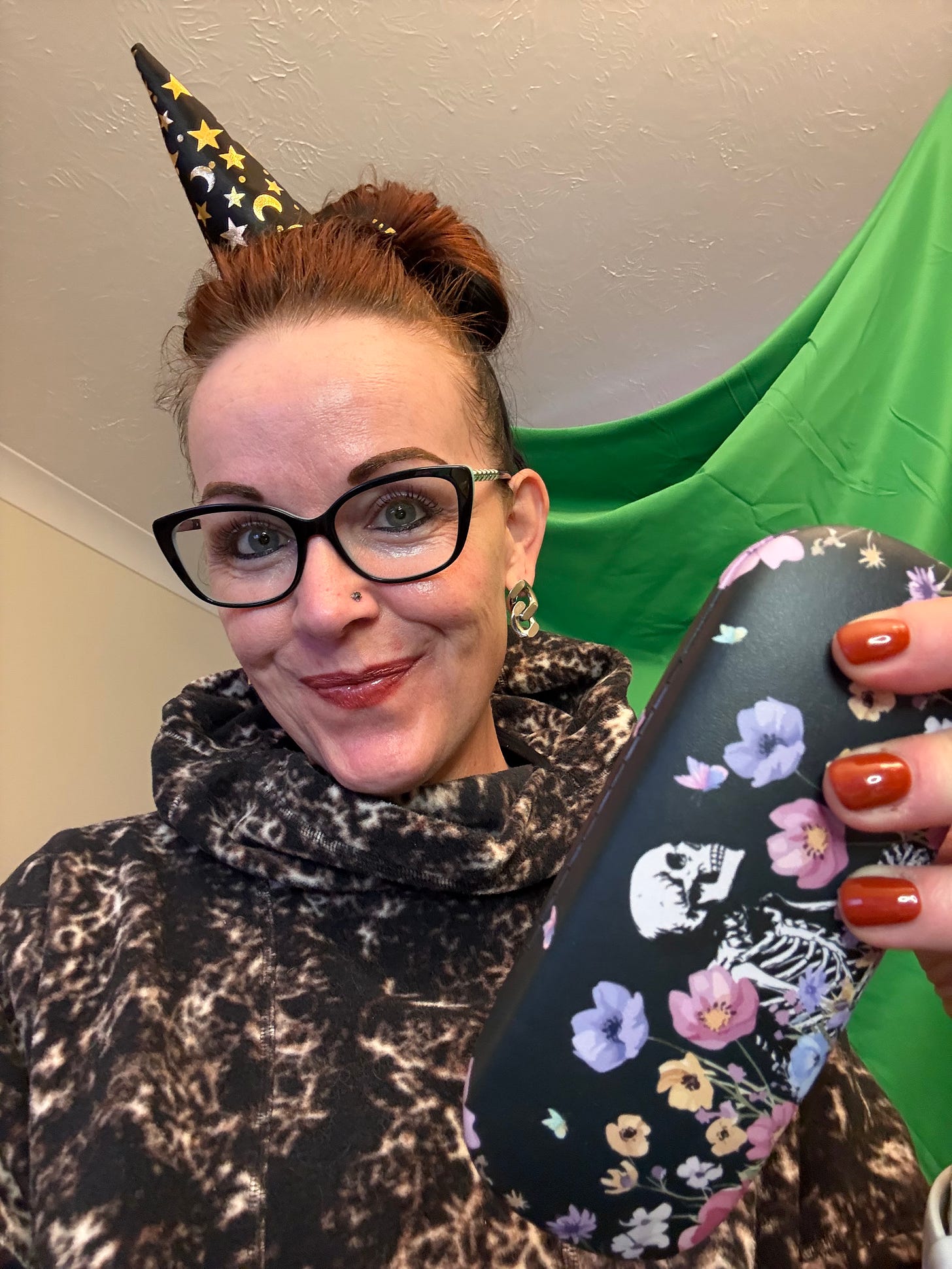 Georgina Dean, wearing a party hat and a leopard-print hoodie, smiles as she holds up her new glasses case, which is black with a floral pattern and a whimsical skeleton.