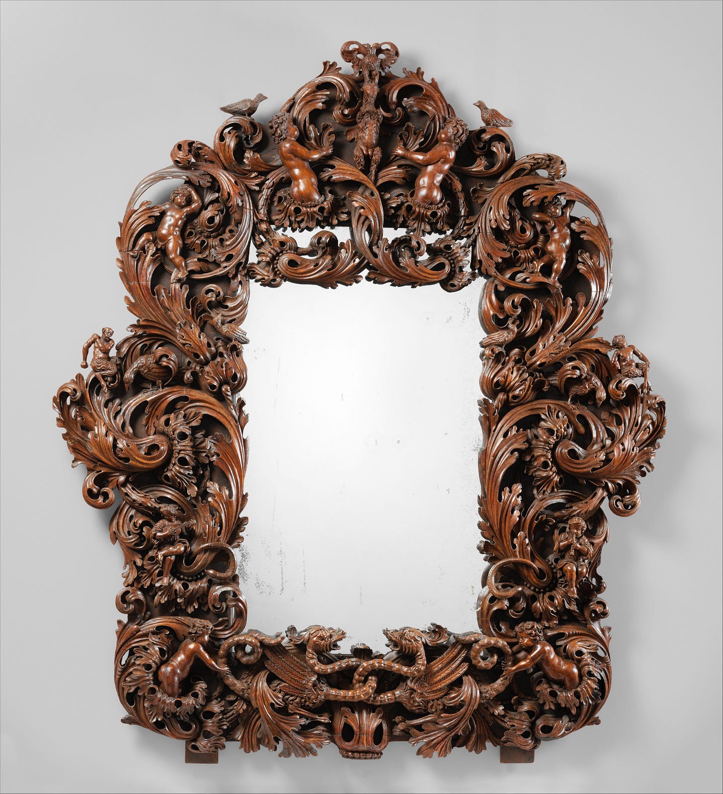 A large, ornately carved and polished wooden mirror. There are figures carved into it such as satyr-like beings and cherub-like figures.