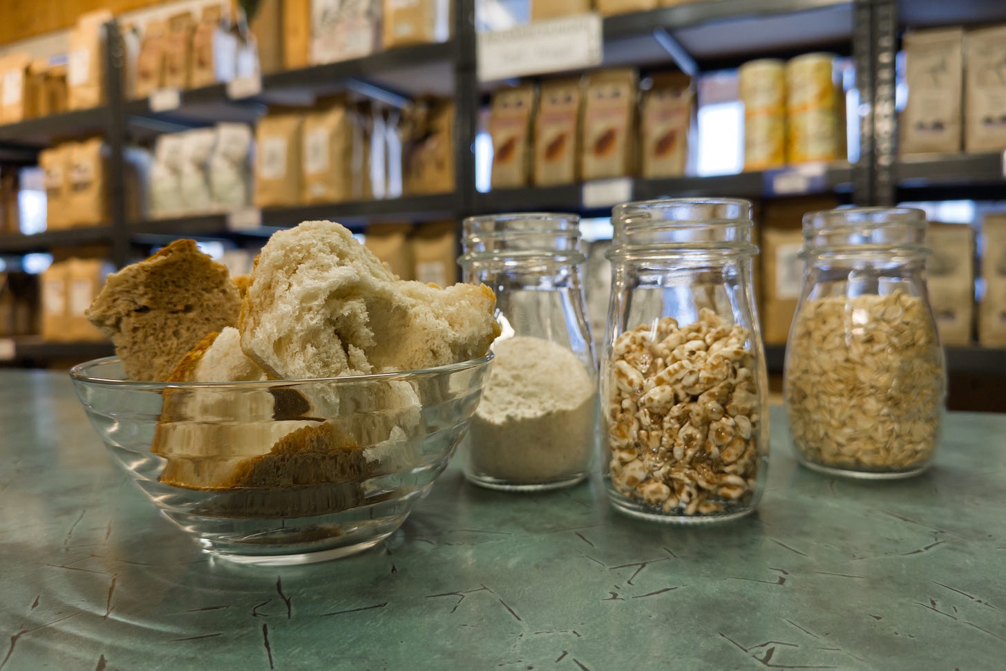 Eine Glasschale mit Restbrot-Stücken steht auf einem grünen Tisch, daneben vier Einmachgläser mit verschiedenen Zutaten: Mehl, Getreideflocken und Körner. Im Hintergrund sind Regale mit Papiersäcken und Lebensmitteln im Laden der Dittligmühle zu sehen. Eine Glasschale mit Restbrot-Stücken steht auf einem grünen Tisch, daneben vier Einmachgläser mit verschiedenen Zutaten: Mehl, Getreideflocken und Körner. Im Hintergrund sind Regale mit Papiersäcken und Lebensmitteln im Laden der Dittligmühle zu sehen.