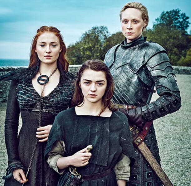 Three female characters from Game of Thrones stand together outdoors, facing the camera with serious expressions. On the left is a red-haired woman in a dark feathered gown with a metal brooch (Sansa Stark). In the center is a shorter, younger woman with dark hair (Arya Stark), wearing a rugged tunic and holding a small weapon. On the right is a tall blonde woman in full armor (Brienne), resting one hand on the hilt of her sword. Behind them is a stone wall and trees under a cloudy sky, evoking a medieval fantasy setting. Three female characters from Game of Thrones stand together outdoors, facing the camera with serious expressions. On the left is a red-haired woman in a dark feathered gown with a metal brooch (Sansa Stark). In the center is a shorter, younger woman with dark hair (Arya Stark), wearing a rugged tunic and holding a small weapon. On the right is a tall blonde woman in full armor (Brienne), resting one hand on the hilt of her sword. Behind them is a stone wall and trees under a cloudy sky, evoking a medieval fantasy setting.