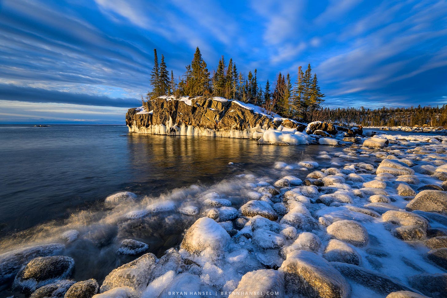 Golden light on an icy rock shoreline leading to an island under stripped clouds in the sky.