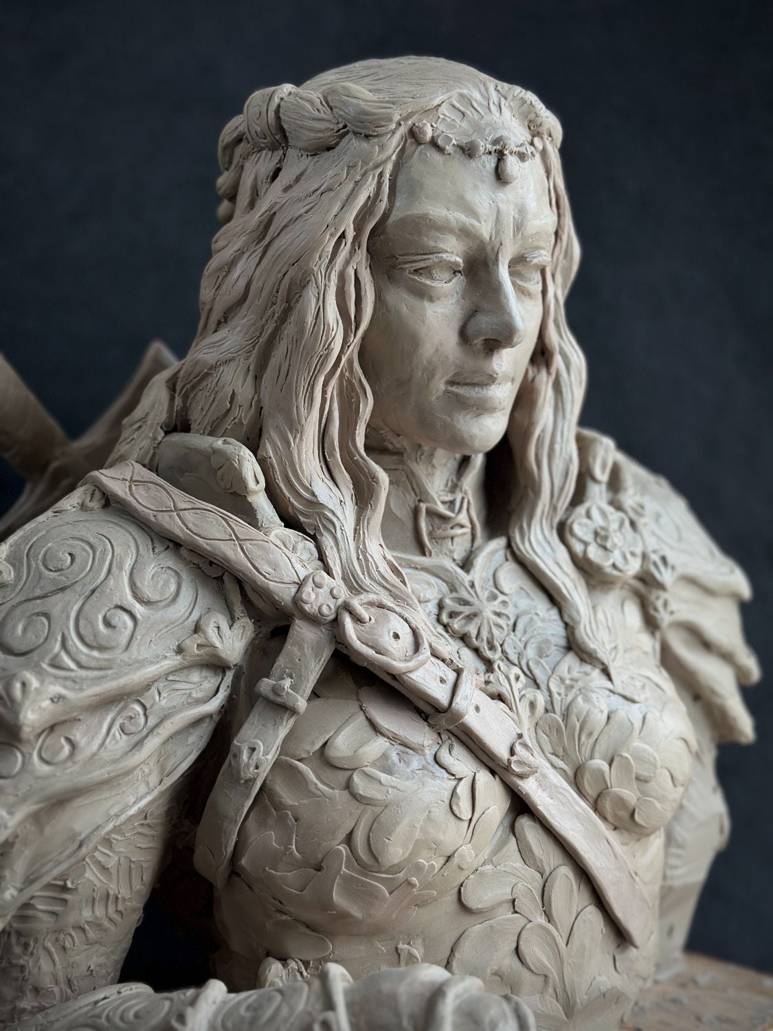 Clay sculpture of an armored warrior woman with long hair, detailed floral patterns, and a sword on a dark background. Mood is serious and focused. Artwork by Kindrie Grove