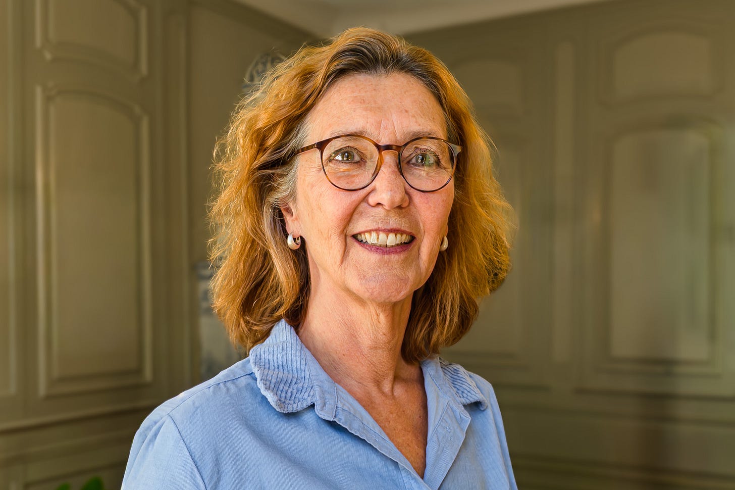 Porträt von Eva Wyss mit schulterlangem, rötlich-braunem Haar und runder Brille. Sie trägt eine hellblaue Bluse und steht vor der dezent getäfelten Wand eines historischen Innenraums. Porträt von Eva Wyss mit schulterlangem, rötlich-braunem Haar und runder Brille. Sie trägt eine hellblaue Bluse und steht vor der dezent getäfelten Wand eines historischen Innenraums.